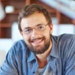A smiling man with glasses and a beard in a casual blue shirt indoors.